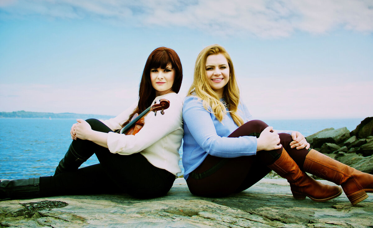 Musical duo portrait Cassie And Maggie sitting backs against each other ocean behind