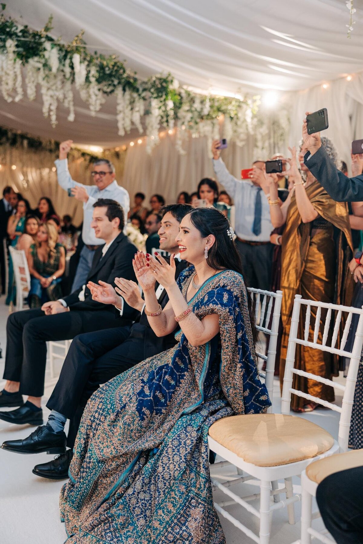 Guests in formal attire clapping and capturing a moment at a wedding reception inside a tent decorated with white drapes and floral arrangements.
