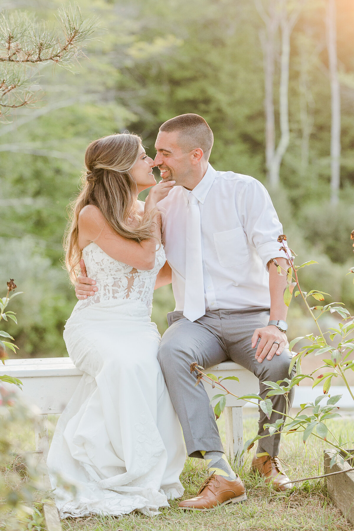 Bride and groom sneak away for a moment at their wedding reception. They are sitting on a bench at the Five Bridge Inn. The bride is lifting the groom's chin and bring him in for a kiss as they are smiling. Captured by best Massachusetts wedding photographer Lia Rose Weddings.