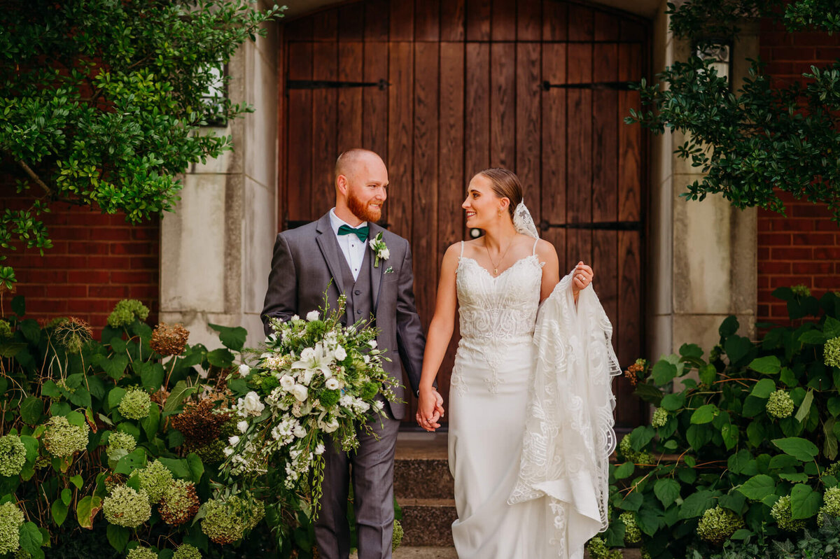 Photo of a bride and groom holding hands and looking at each other in front of a wooden door with greenery in the background