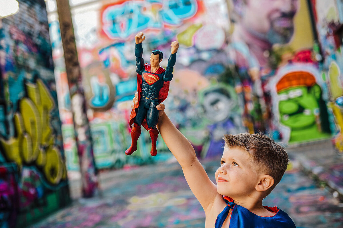 Little boy in blue superhero cape playing with  a superman action figure in graffiti alley near MICA in Baltimore Maryland