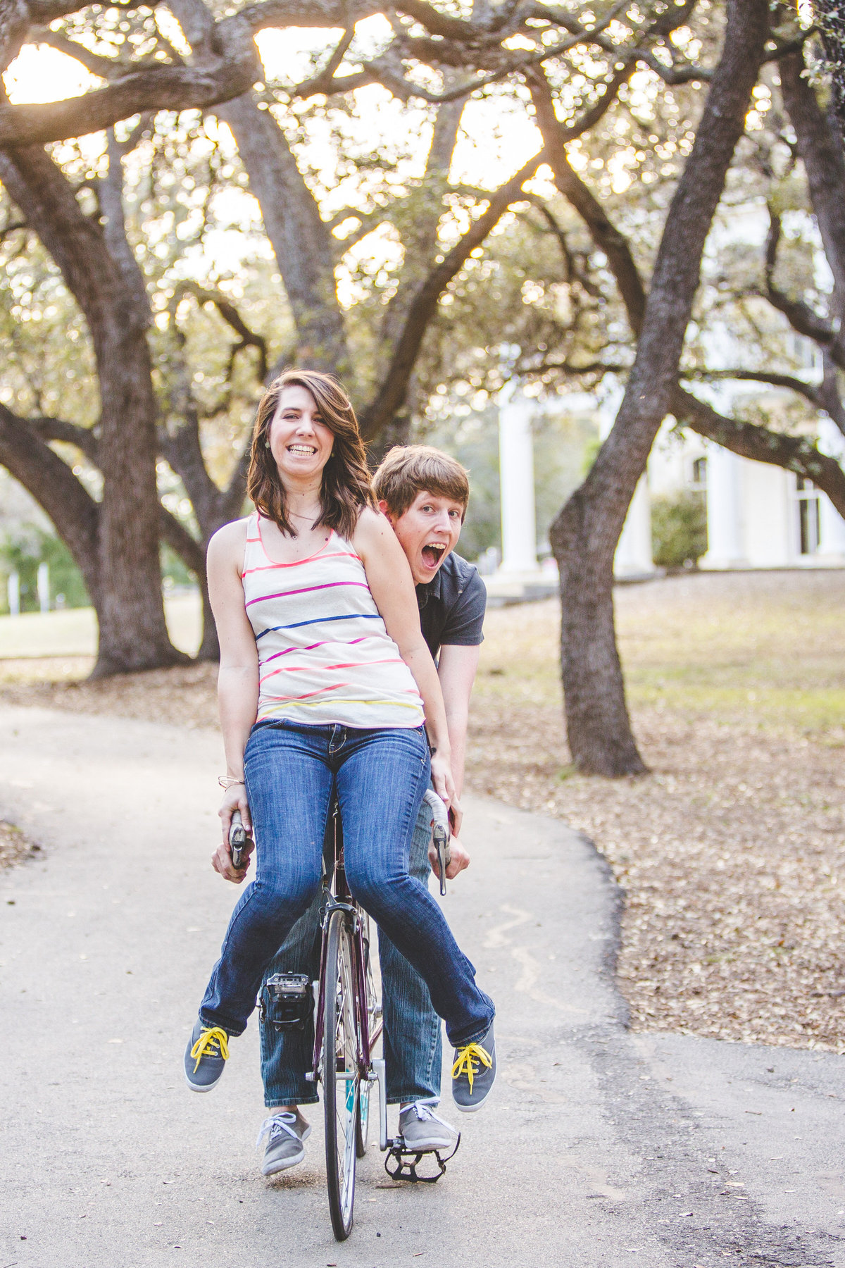 Engaged couple riding one bike together. Fiancée is on the handlebars and fiancé is pedaling at Denman Estate Park in San Antonio.