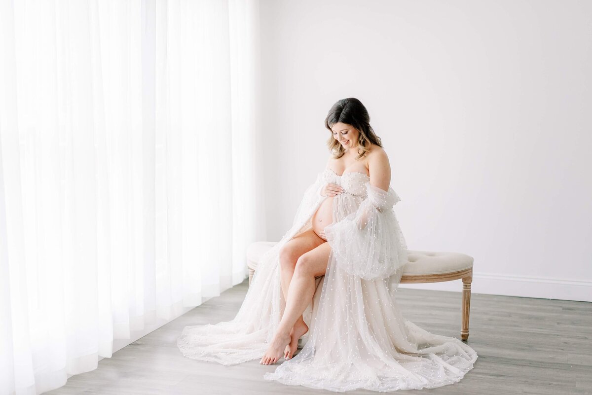 Mom in a big tulle gown elegantly poses on a studio settee  with her toes pointed out and hands holding her exposed baby bump.  Captured by Melissa Mayrie Photography, a maternity photographer in Charlotte, NC.