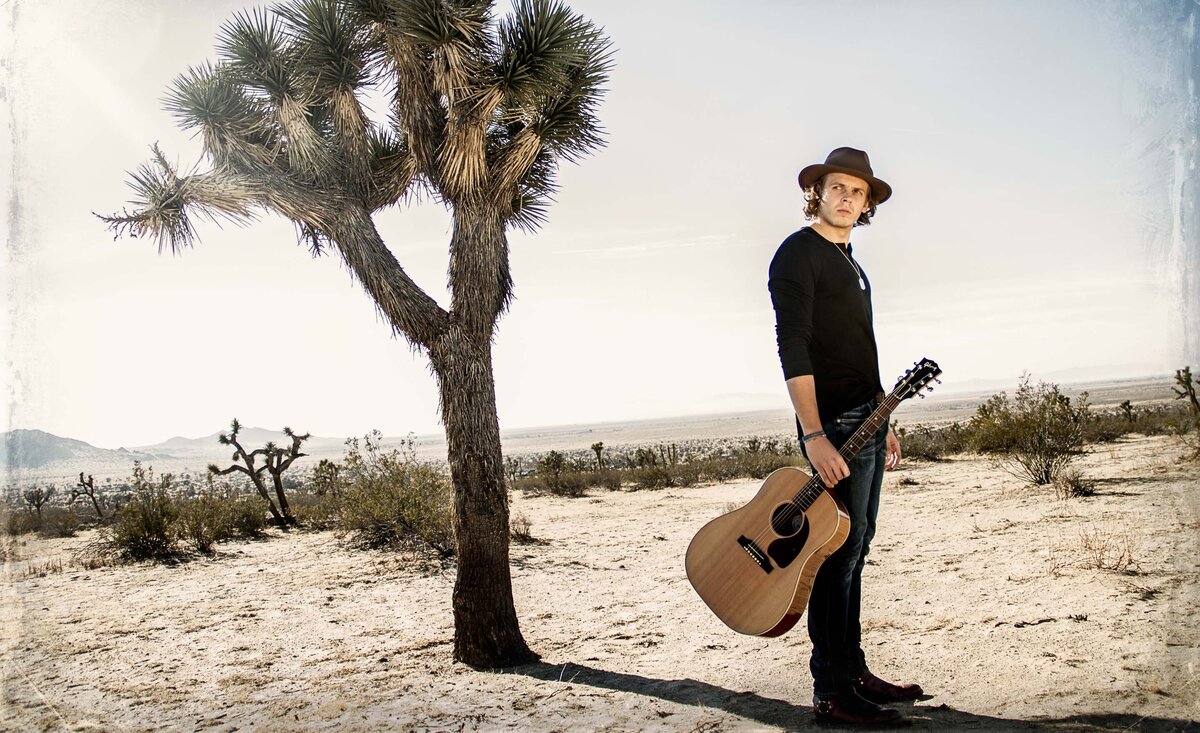 Country music photo Cole Bradley holding guitar wearing black shirt cowboy hat beside Joshua Tree desert background