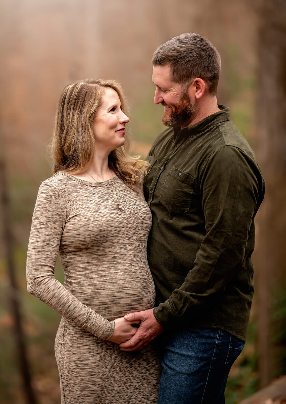 An expecting couple  hugs and laughs while standing in the forest in Asheville