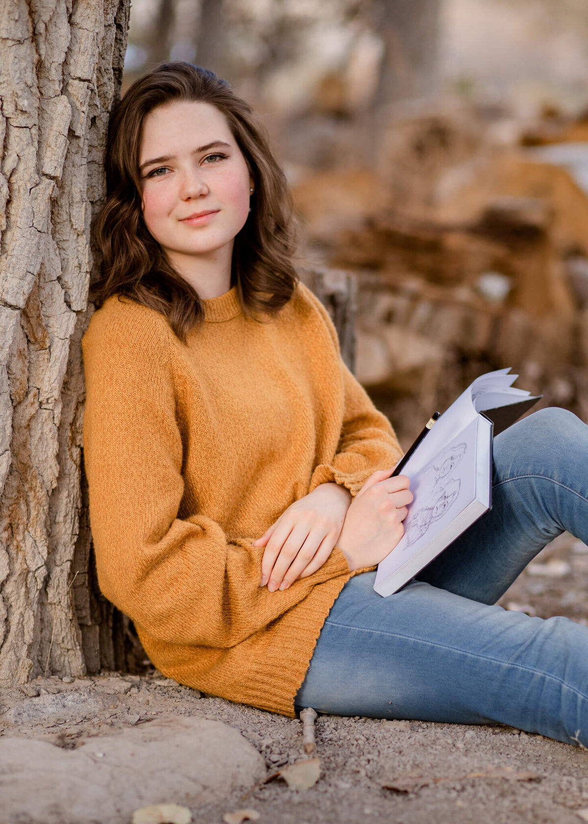 teenage girl sitting leaning against a tree holding her sketch book and pencil