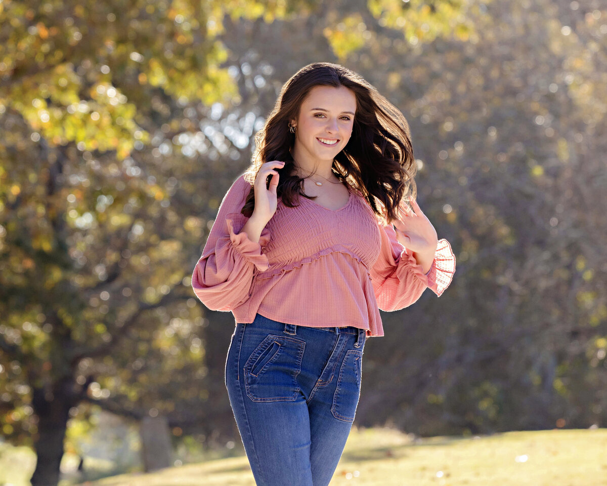 HS senior girl in pink blouse and jeans smiling on gravel path surrounded by golden leaves at Anne Springs Close Greenway