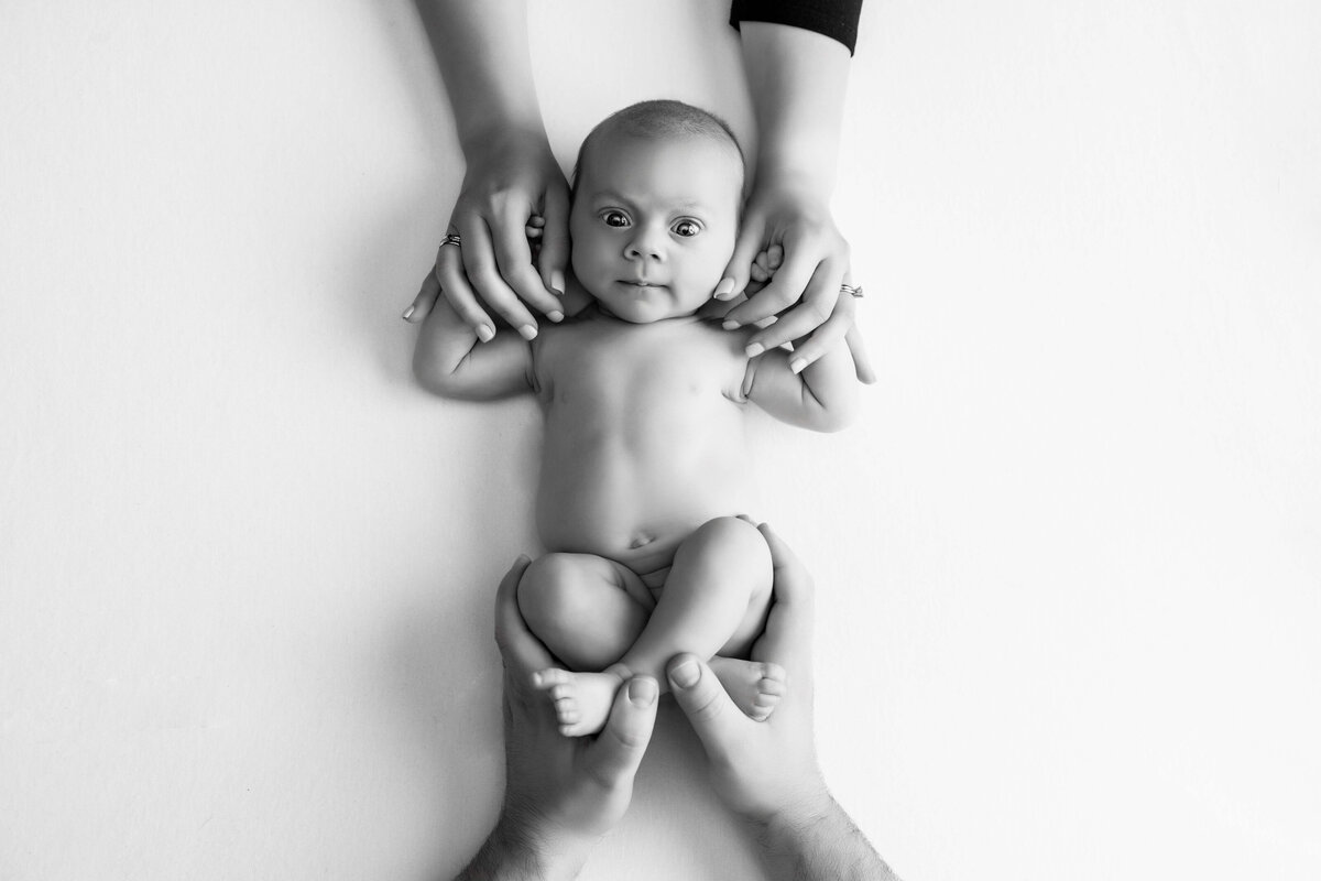 Black and white image of a baby lying on a white surface, looking up. The baby's hands are gently held by an adult's hands at the top, and its feet are cradled by two adult hands at the bottom—a signature capture by a Jacksonville baby photographer.