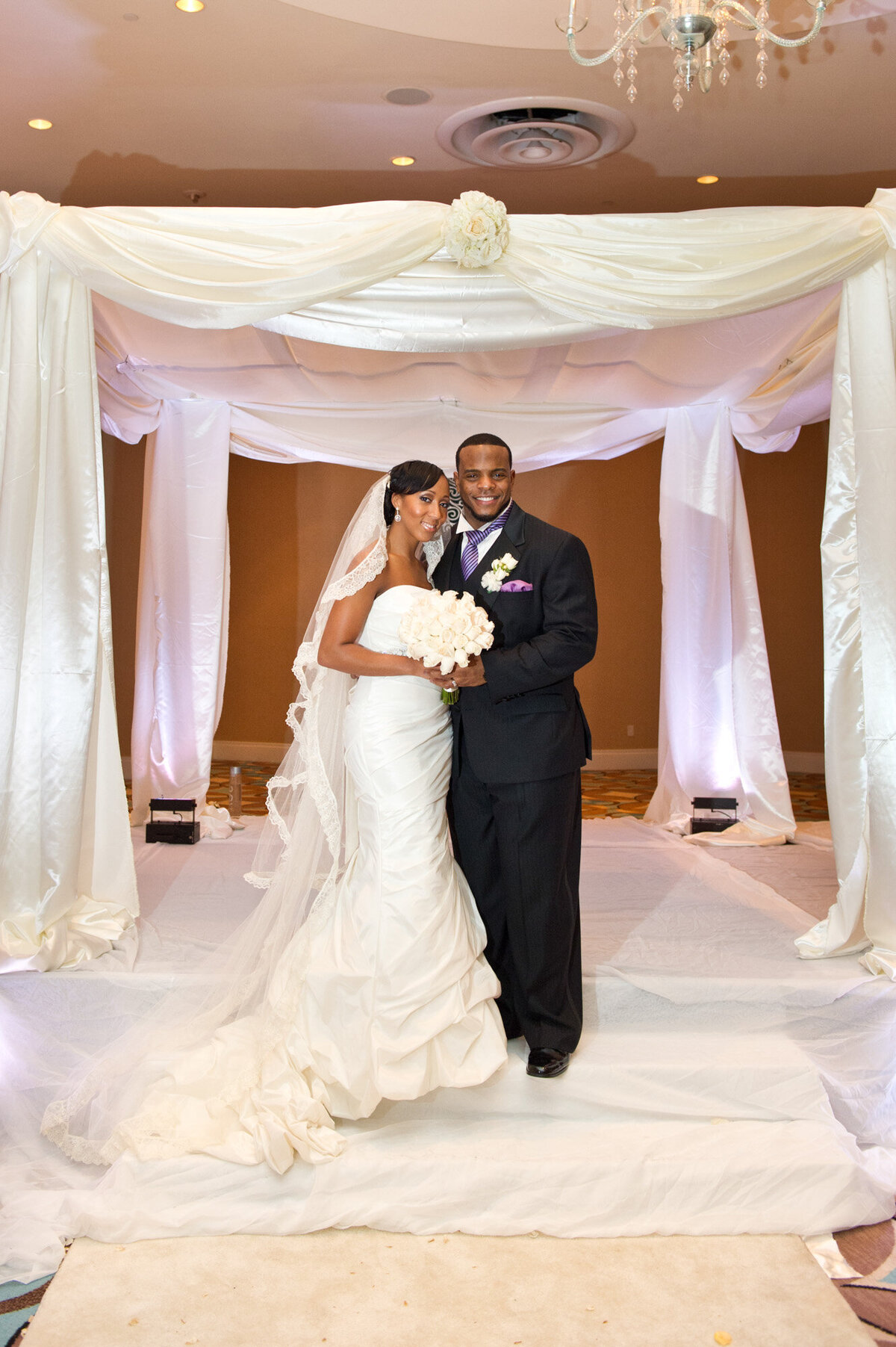 Newly wedded couple holding white floral bouquet with white draping behind