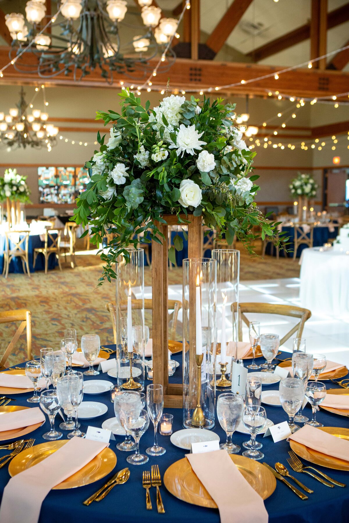 Elegant wedding table setting at a park farm winery, featuring a tall floral centerpiece, navy blue tablecloth, gold accents, and glassware under string lights.