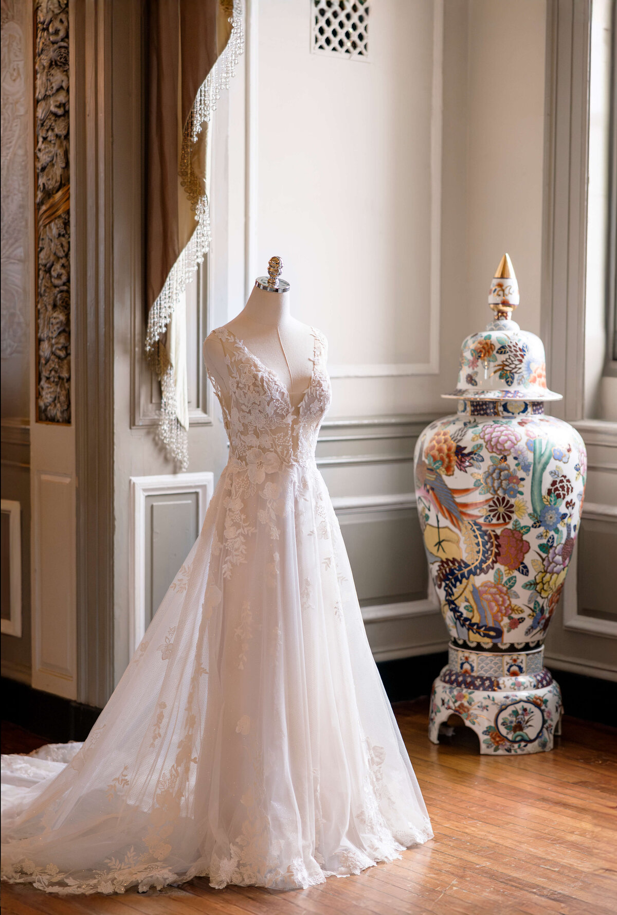 A classic white wedding dress with floral lace details is displayed on a mannequin in a luxurious room. Next to it is a large ornate vase with intricate patterns, set against an elaborately decorated wall.