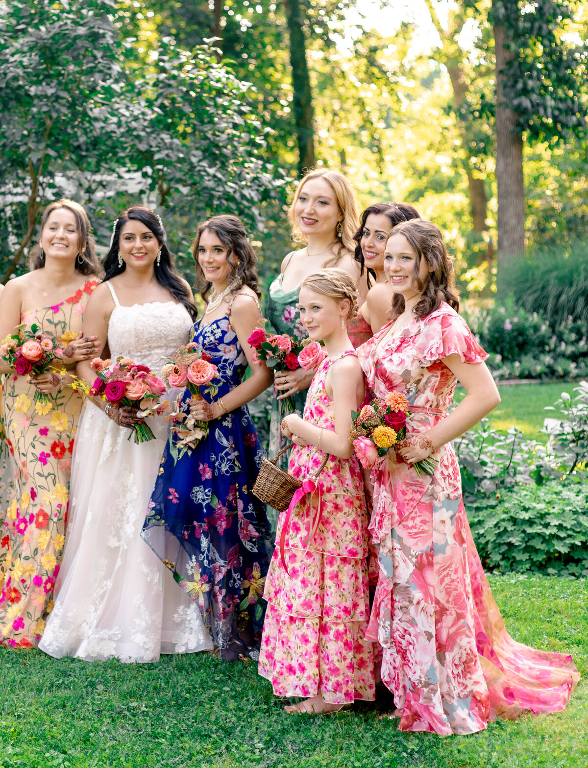 A group of women wearing colorful floral dresses stand together in a lush garden, holding bouquets of flowers. The setting is bright and outdoors, with tall trees and greenery in the background.