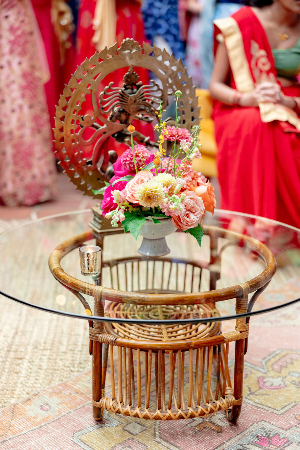 A glass-topped rattan table holds a pot of vibrant flowers, including pink and orange roses. A decorative metal sculpture is in the background, with people in colorful traditional attire partially visible.