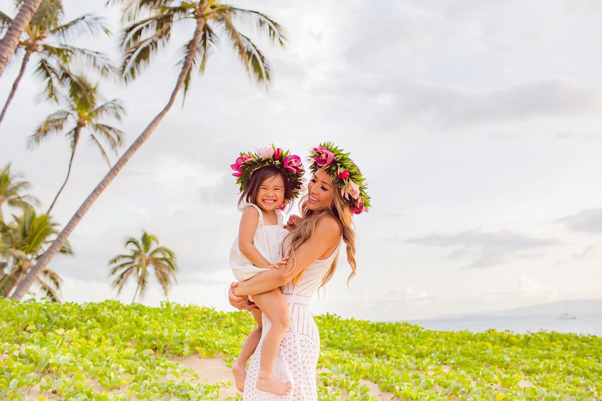 Beautiful brunette mom holds toddler on her hips and wears a vibrant floral crown during their family photography session at the beach