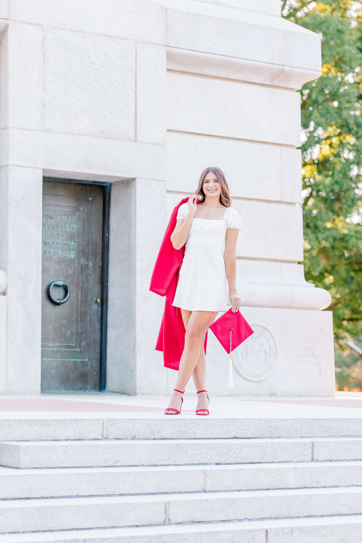 A person in a white dress stands on stone steps, holding a red graduation cap and gown over their shoulder. Captured by a talented senior portrait photographer in Fayetteville, NC, they are smiling amidst the backdrop of trees and a large stone building.