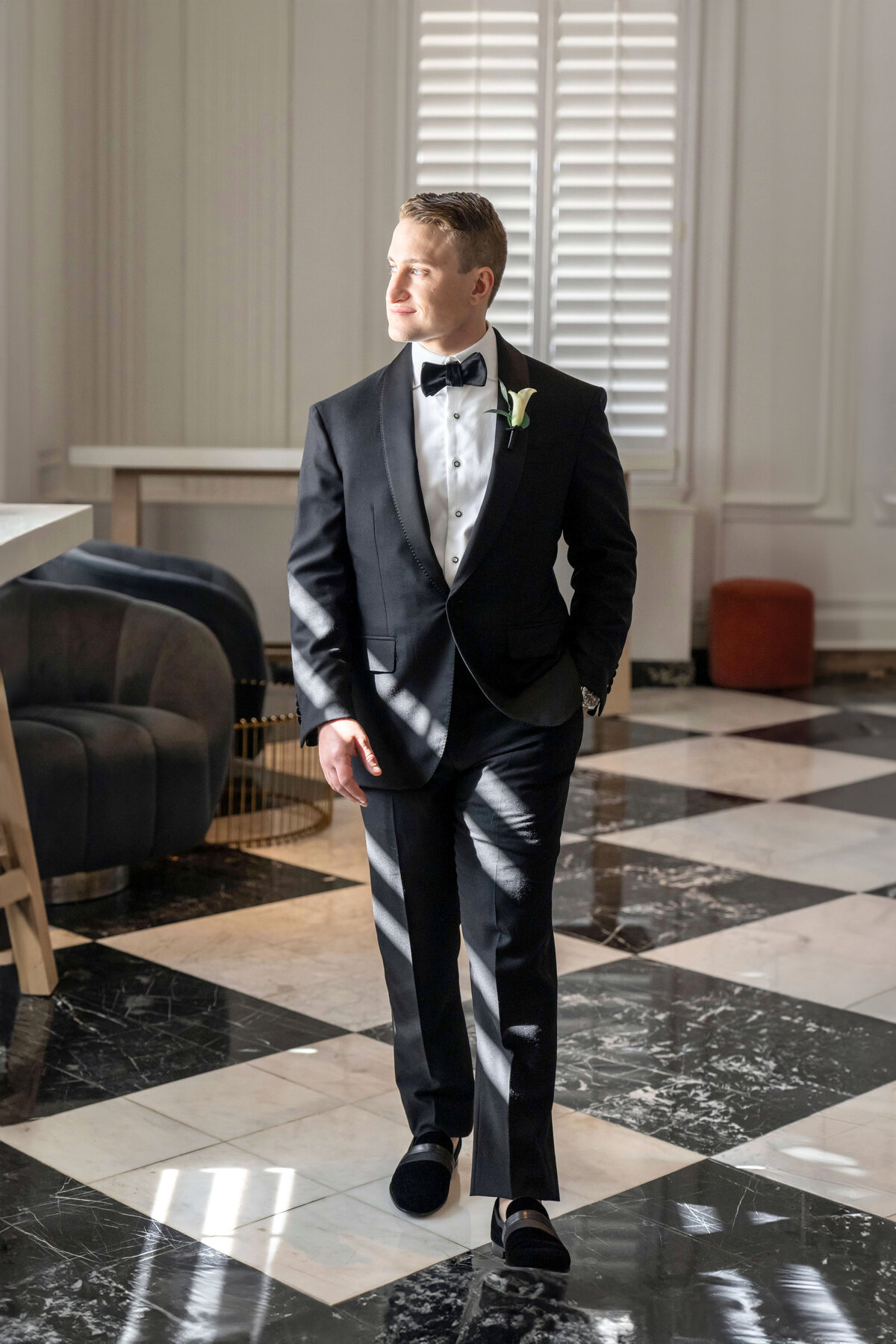 A man in a black tuxedo and bow tie walks confidently across a marble floor in a sunlit room with white walls and tall shutters. He has a boutonniere on his lapel and is smiling slightly.