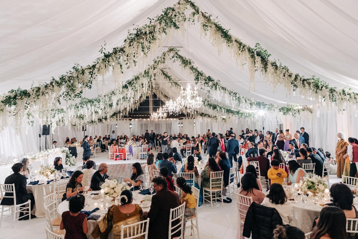 Elegant wedding reception in a tent adorned with floral garlands and chandeliers, with guests seated at tables.