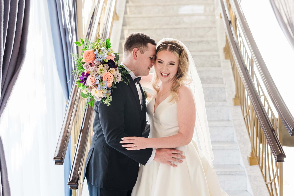 A bride and groom smiling and embracing on a staircase, with the bride holding a bouquet of colorful flowers, at an event in Davenport.