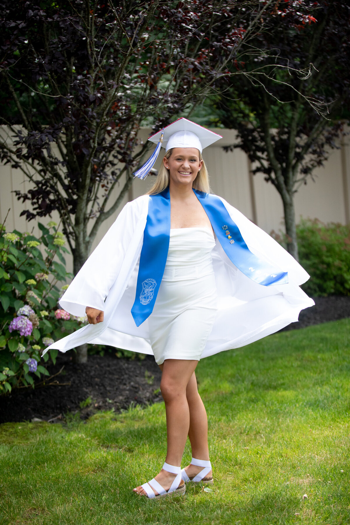 white teenage girl with blonde hair twirls in a white graduation cap and gown with a blue stole Pearl River senior portrait photographer Milestone Images