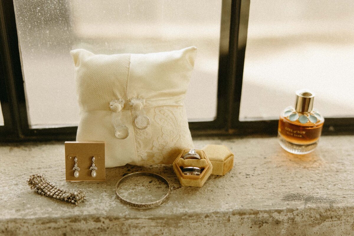 Iowa weddings accessories on a windowsill, including a ring pillow, earrings, a bracelet, a perfume bottle, and a ring box.