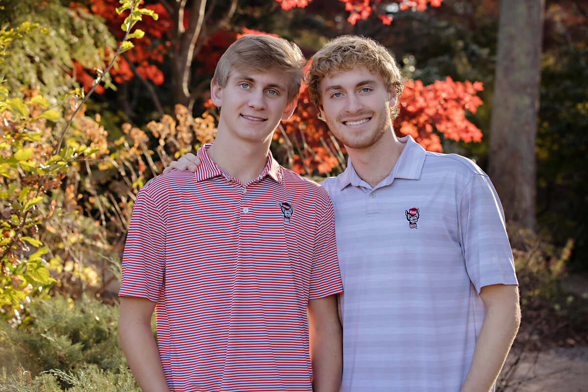 Striking autumn sibling portrait of two red‑haired brothers in NC State University shirts and sweaters surrounded by golden foliage at UNCC Botanical Garden Charlotte NC