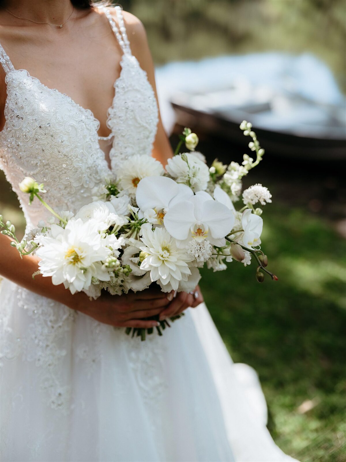 Detailed photo of bridal bouquet of white orchids, sparkly dress, and wooden rowboat in background.
