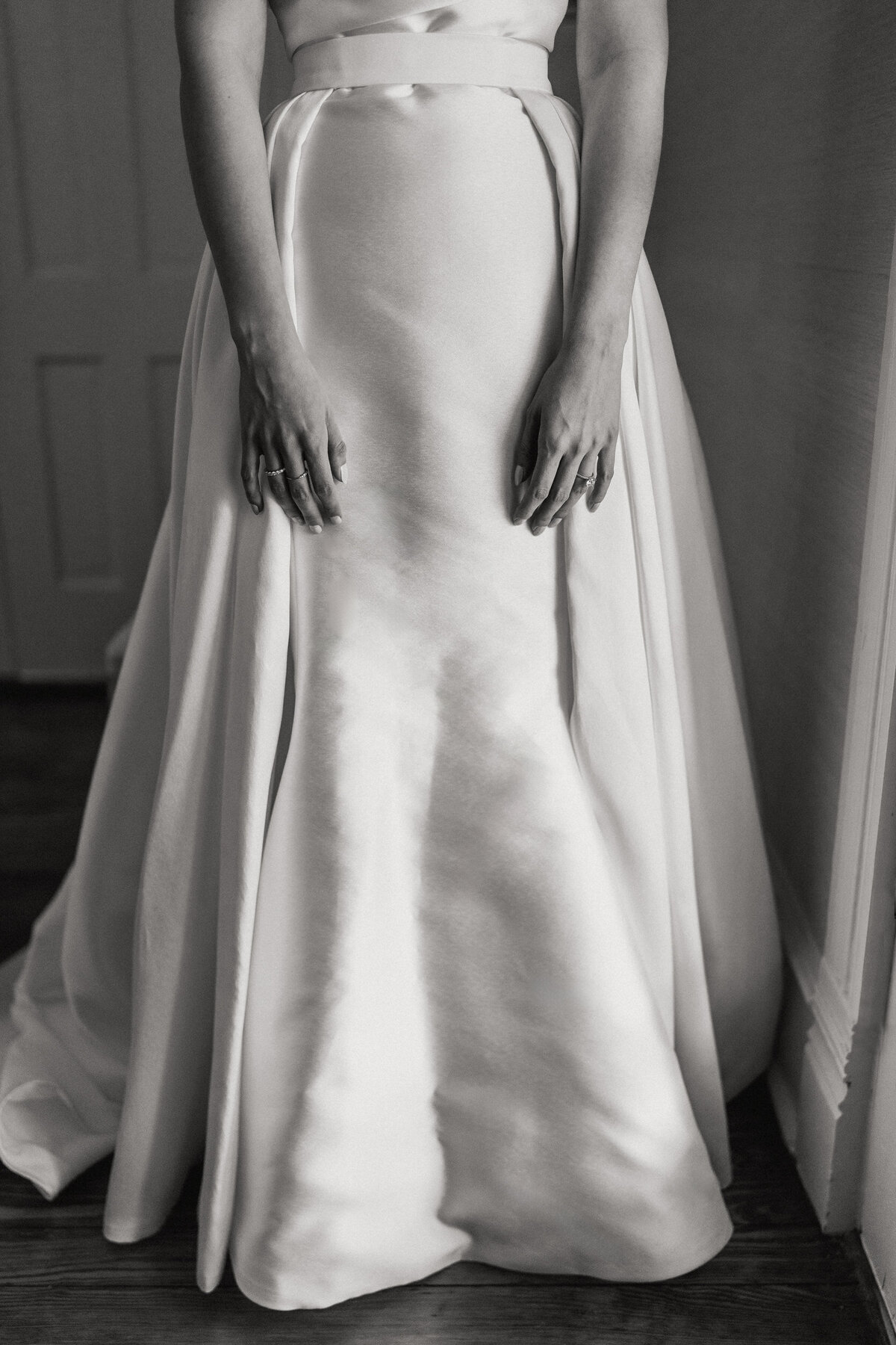 A black and white photograph of Ixi on her wedding day at Woodbine Mansion in Round Rock, near Austin, TX. The photograph is from the waist down showing the satin fabric of the bride’s wedding dress and the additional waist skirt that adds draping on both sides. Her hands are gently resting at her sides and the scene is beautifully lit by window light from the right. Wedding photography by Stacie McChesney/Vitae Weddings.