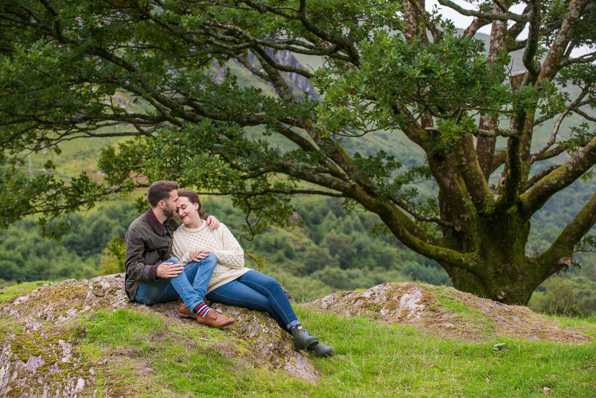 Young engaged couple in aran sweater and denim jeans kissing under a tree in a field