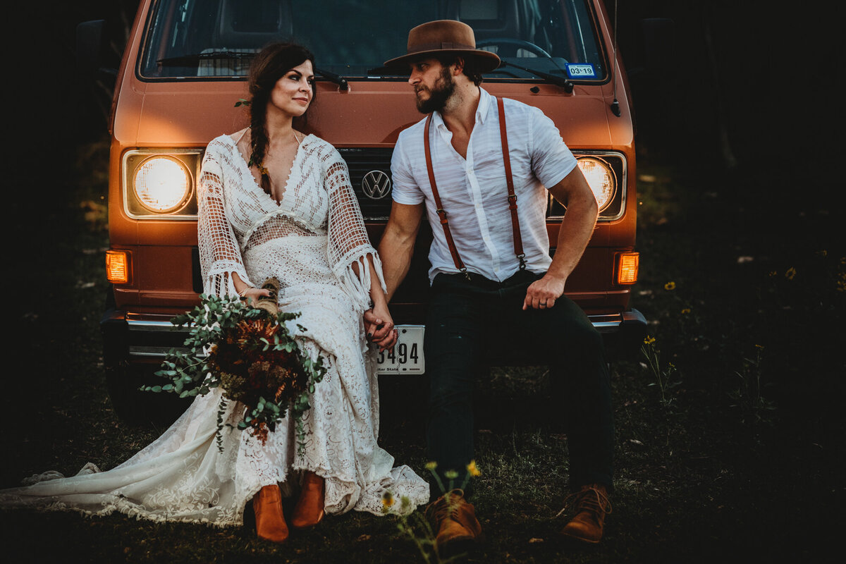 Couples Photography, Man in suspenders and a brown hat holds hands with woman in a vintage lace dress holding a bouquet and look at each other in front of a vintage rust colored VW bus.