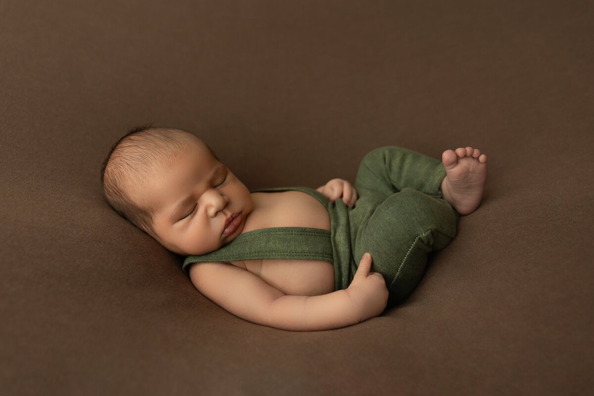 A sleepy newborn baby boy in green overalls sleeps on a brown bed in a studio of a NJ Newborn Photography