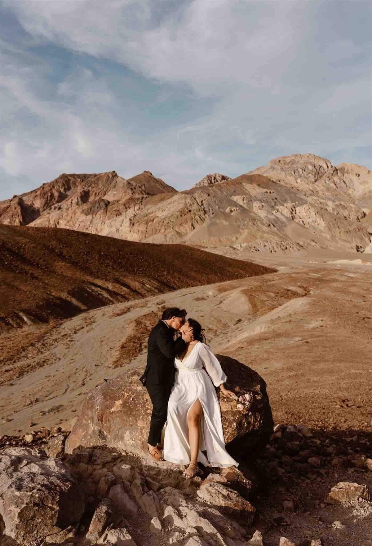 Two brides sit on a rock, sharing a kiss with layered desert mountains in the background.
