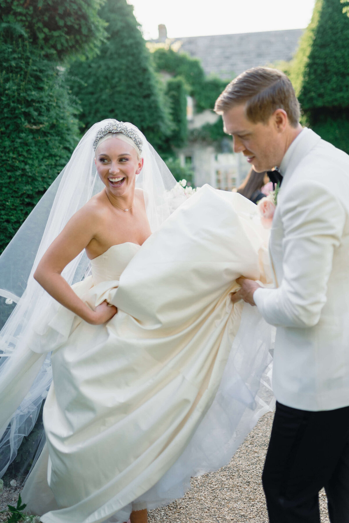 bride laughs as the groom helps her walk by lifting her wedding dress in the gardens of their romantic cotswold wedding venue at euridge manor
