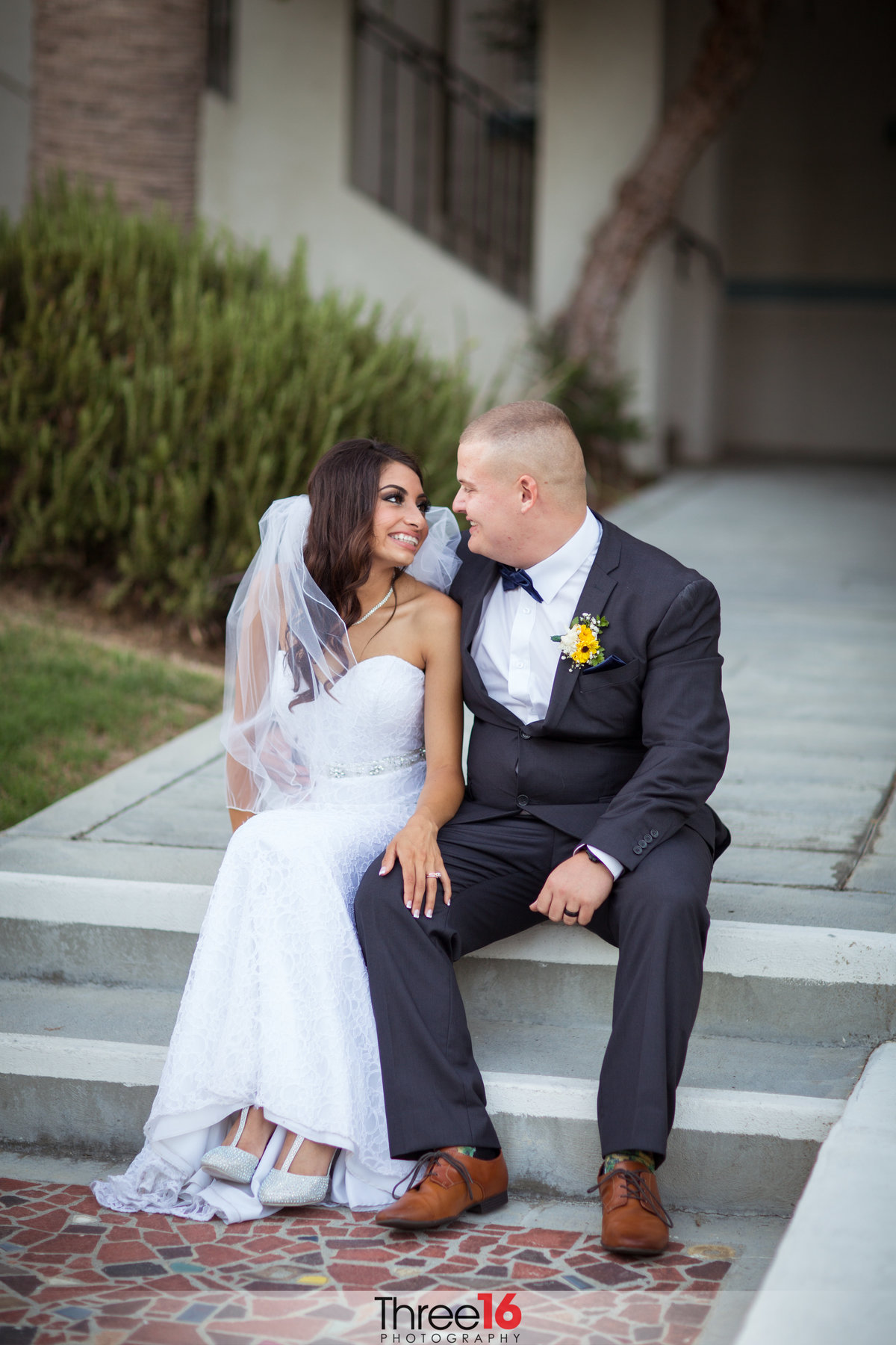 Bride and Groom smile at each other as they sit on a cement step and gaze at each other