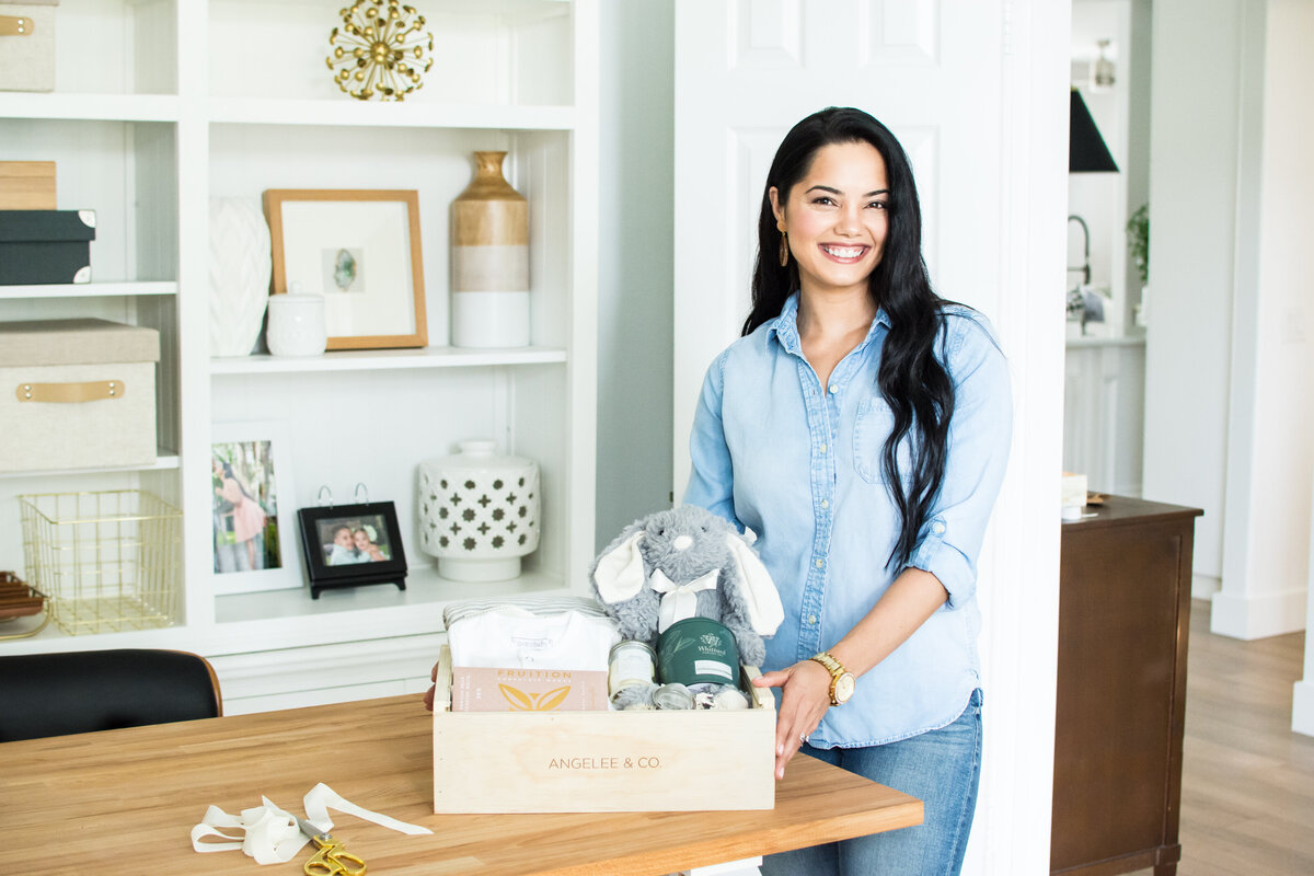 Smiling woman with dark hair in a brightly lit home office showing a curate gift box for a mom and new baby