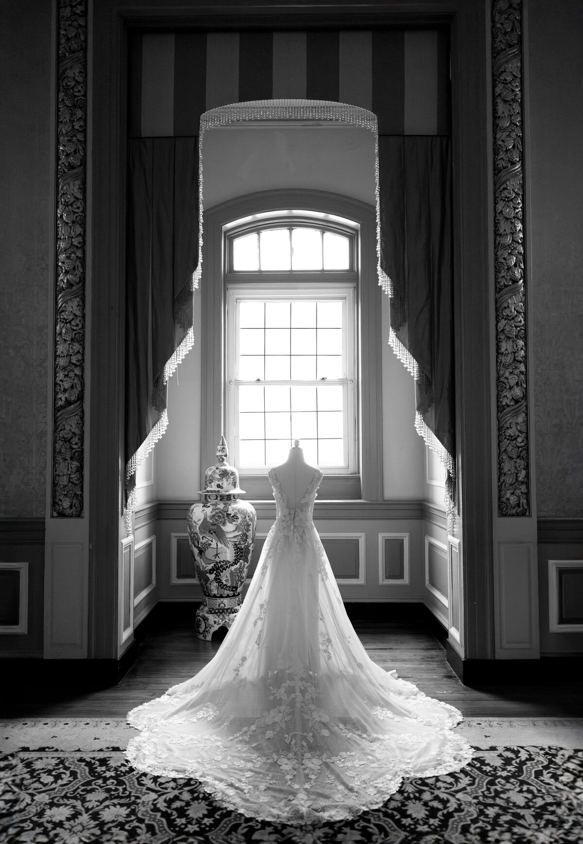 A long, elegant wedding dress on a mannequin is displayed in front of a tall, arched window with draped curtains. The room features intricate paneling and a patterned rug, with a decorative vase placed beside the dress.