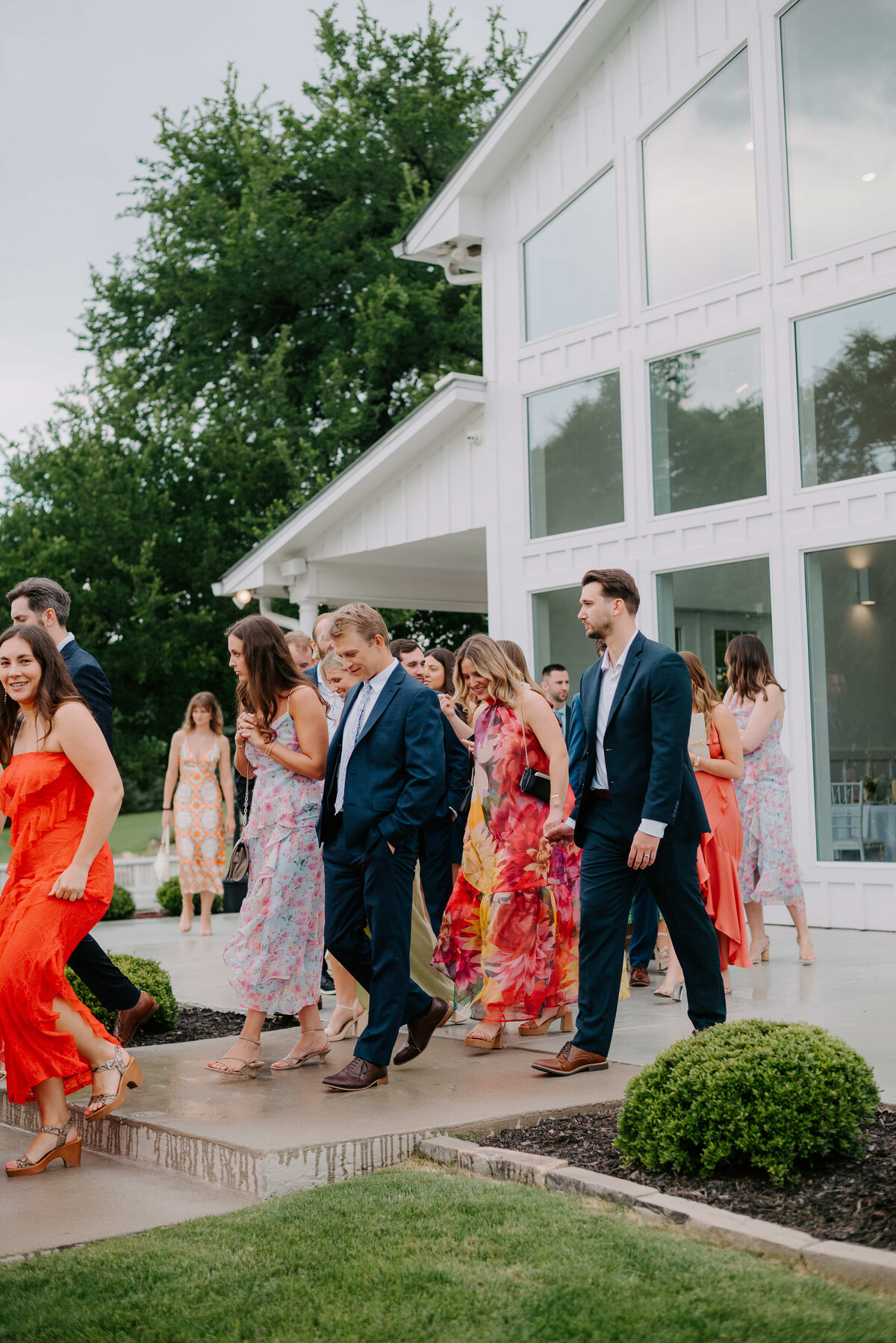 A group of well-dressed people in colorful attire mingles outside a modern white building with large windows. The setting is lively and cheerful.