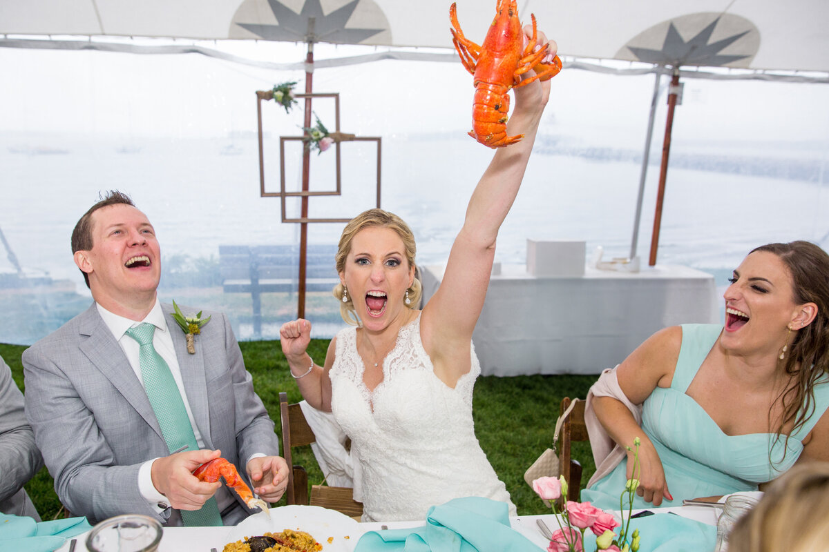 A bride raising up a lobster while sitting at a table with her wedding party.
