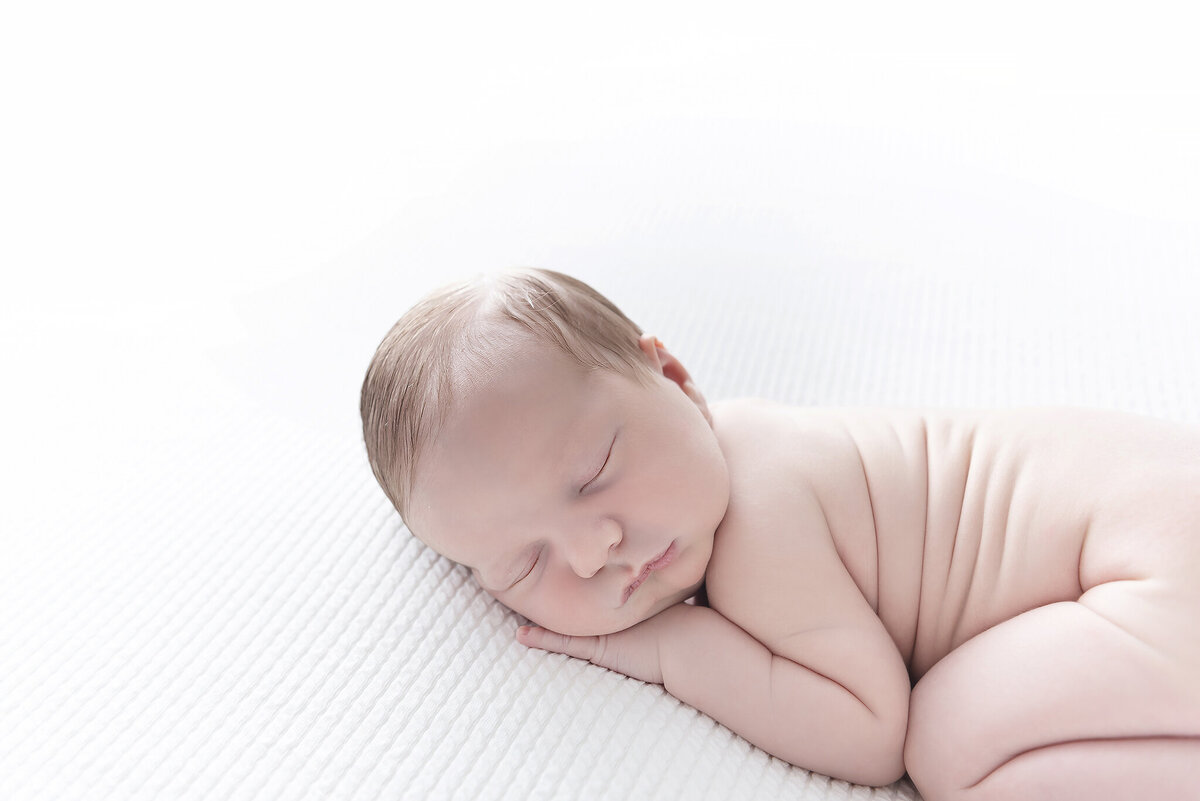 A newborn baby sleeps naked in froggy pose in a studio on a white bed