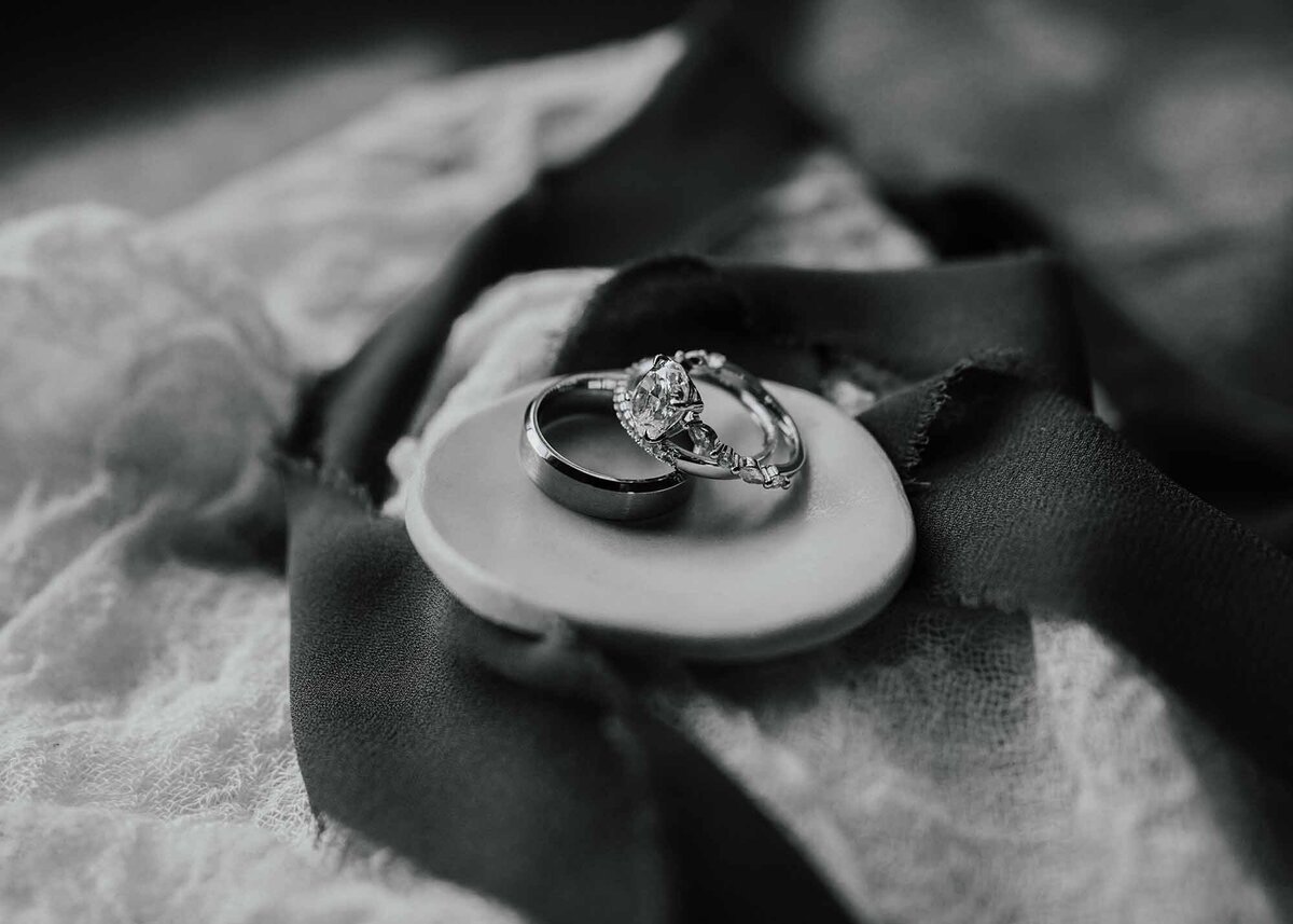 Maddie Rae Photography black and white detail shot of the bride and grooms rings on a little clay bowl. ther eis ribbon and some fabric in the background