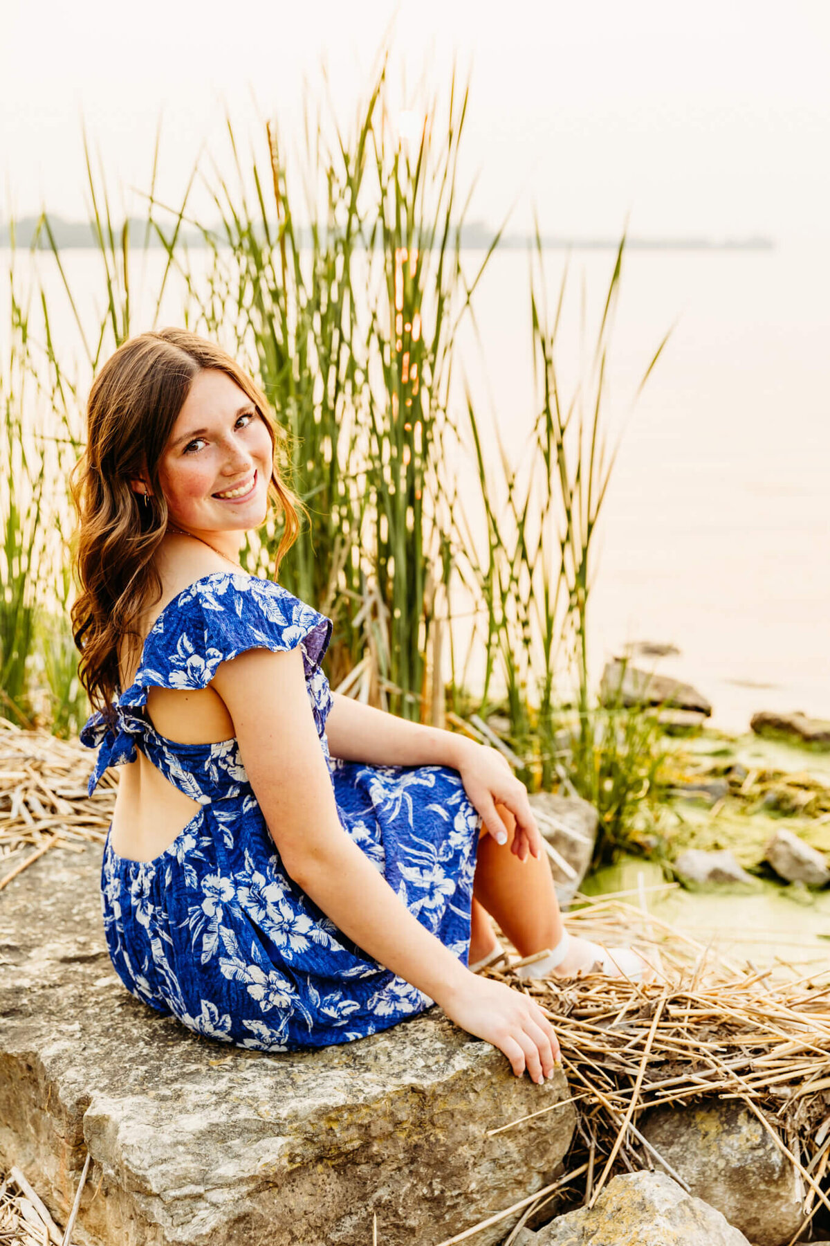teenage girl in a blue dress with white flowers sitting lakeside as she looks over her shoulder while watching the sun set captured by Ashley Kalbus Photography
