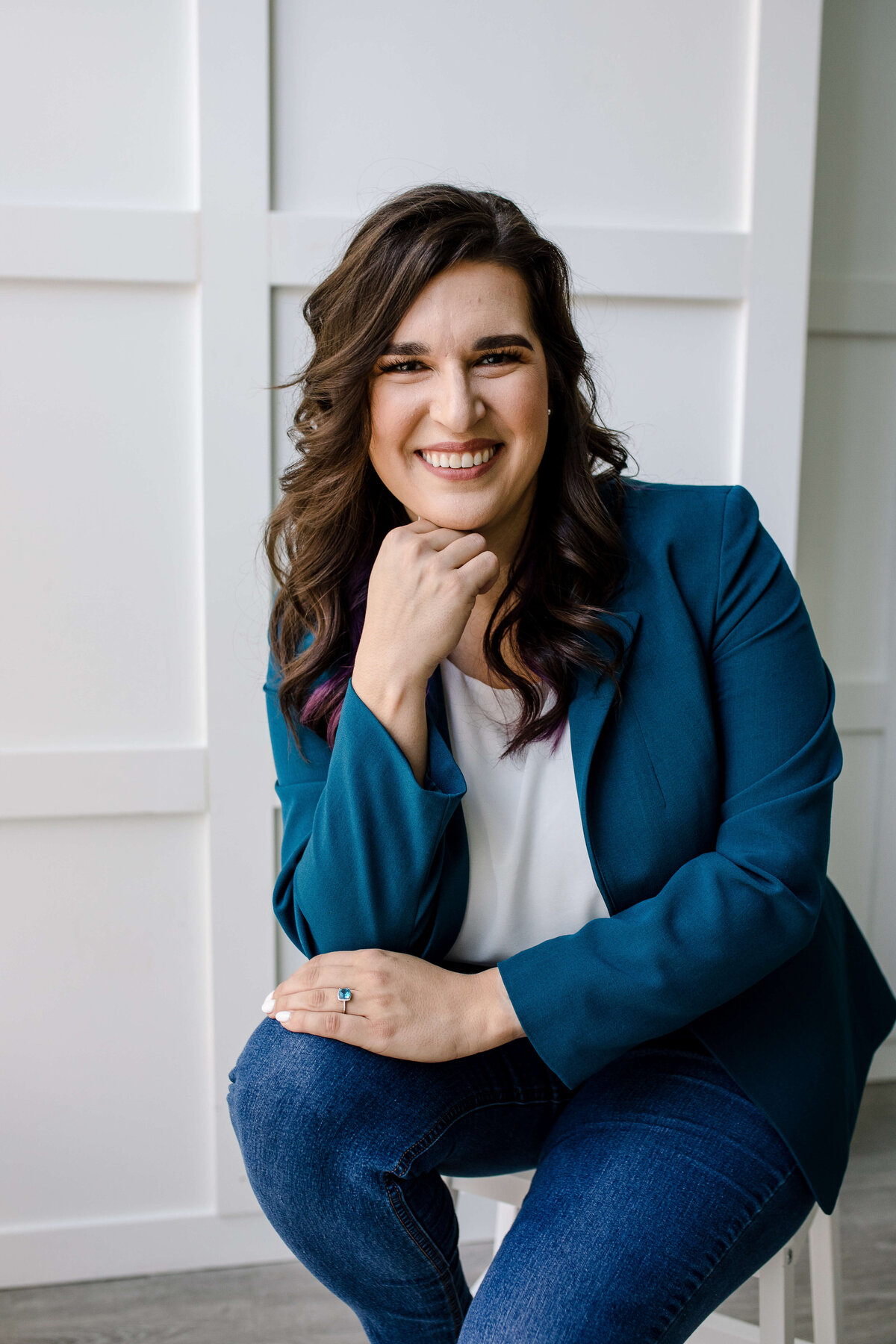 Headshot poses for business owners with woman and blue jeans and a tail blazer sitting on a stool in a studio brand photo shoot with her elbow resting on one of her niece and her other hand resting on her leg as she smiles