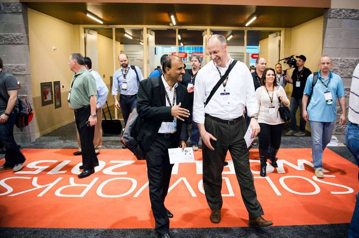Two attendees look toward eachother as they walk in front of a large crowd while entering the expo hall