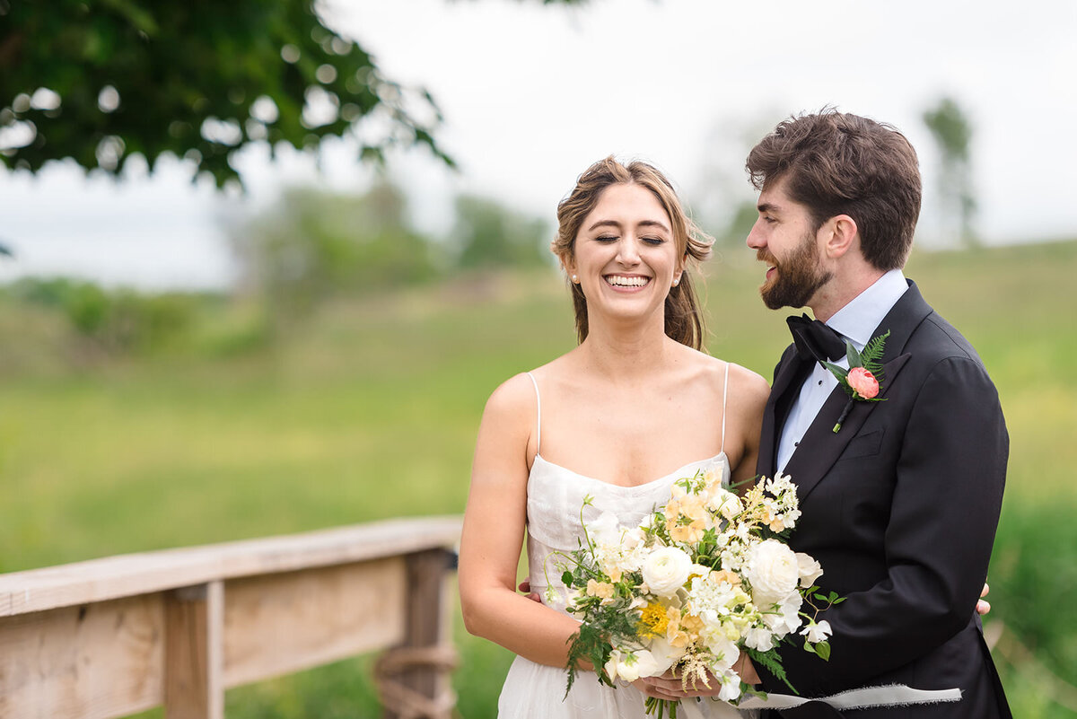 A bride and groom smiling at each other, in an outdoor setting with greenery in the background.