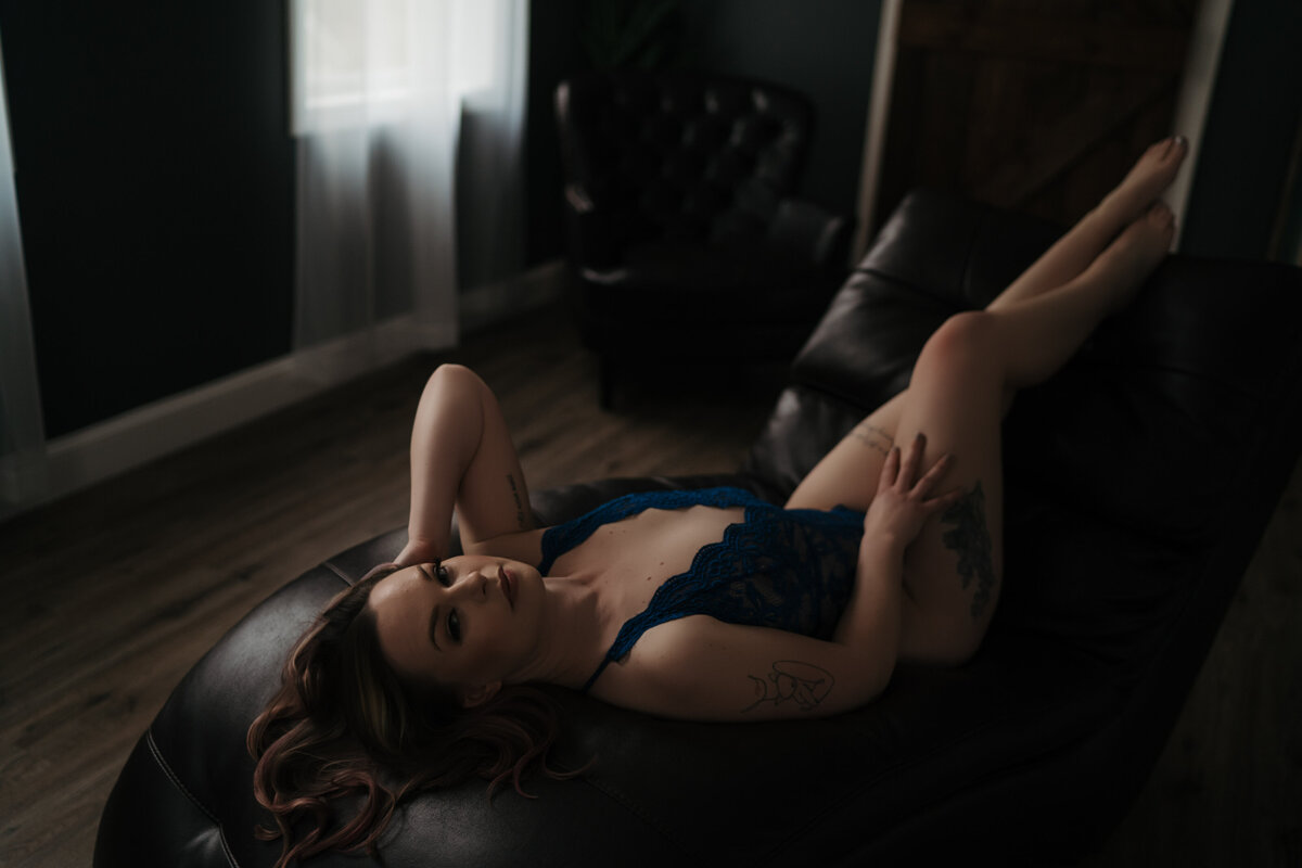 A woman in blue lace lingerie lays on a black leather chaise lounge while running a hand through her hair
