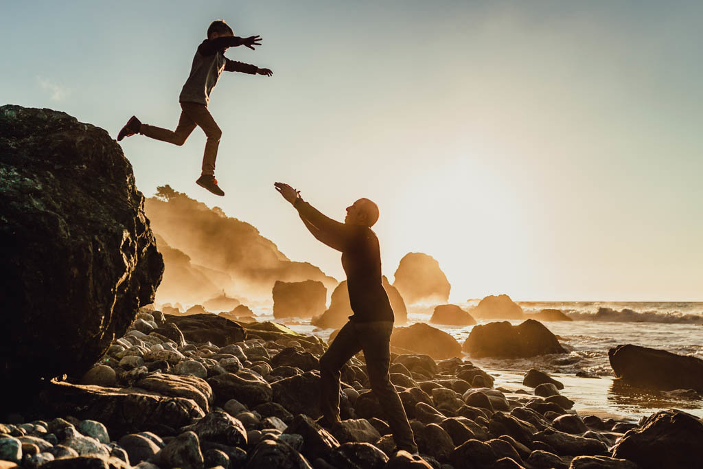 Boy jumps of large rock toward dad with outstretched arms on rocky California Beach
