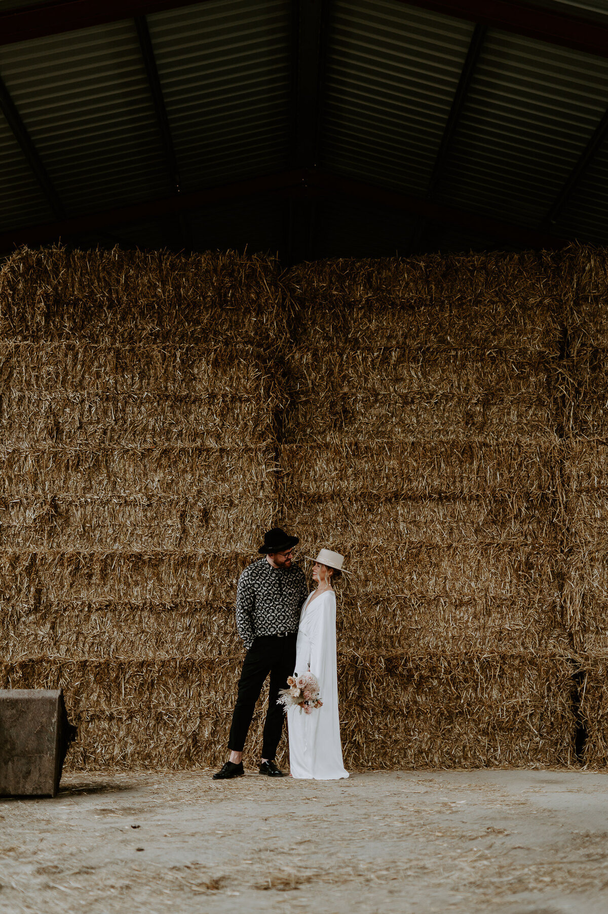 A bride and groom stand together infant of huge hay bales, the bride is wearing an oho wedding dress and a cool hat. The grooms wearing dr Martens, a black and white pattern shirt and matching hat.