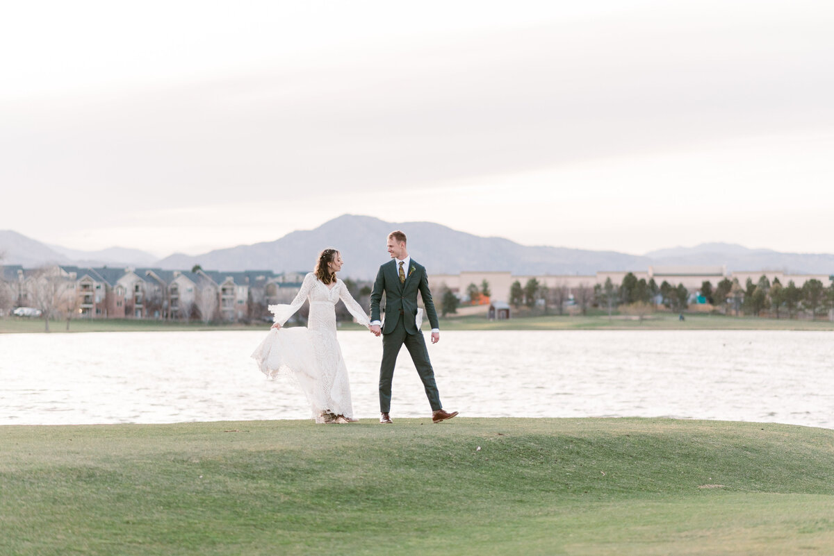 outdoor wedding photos for a denver colorado wedding with bride adn groom holding hands and walking along the side of a lake with the mountains in the distance captured by denver wedding photographer