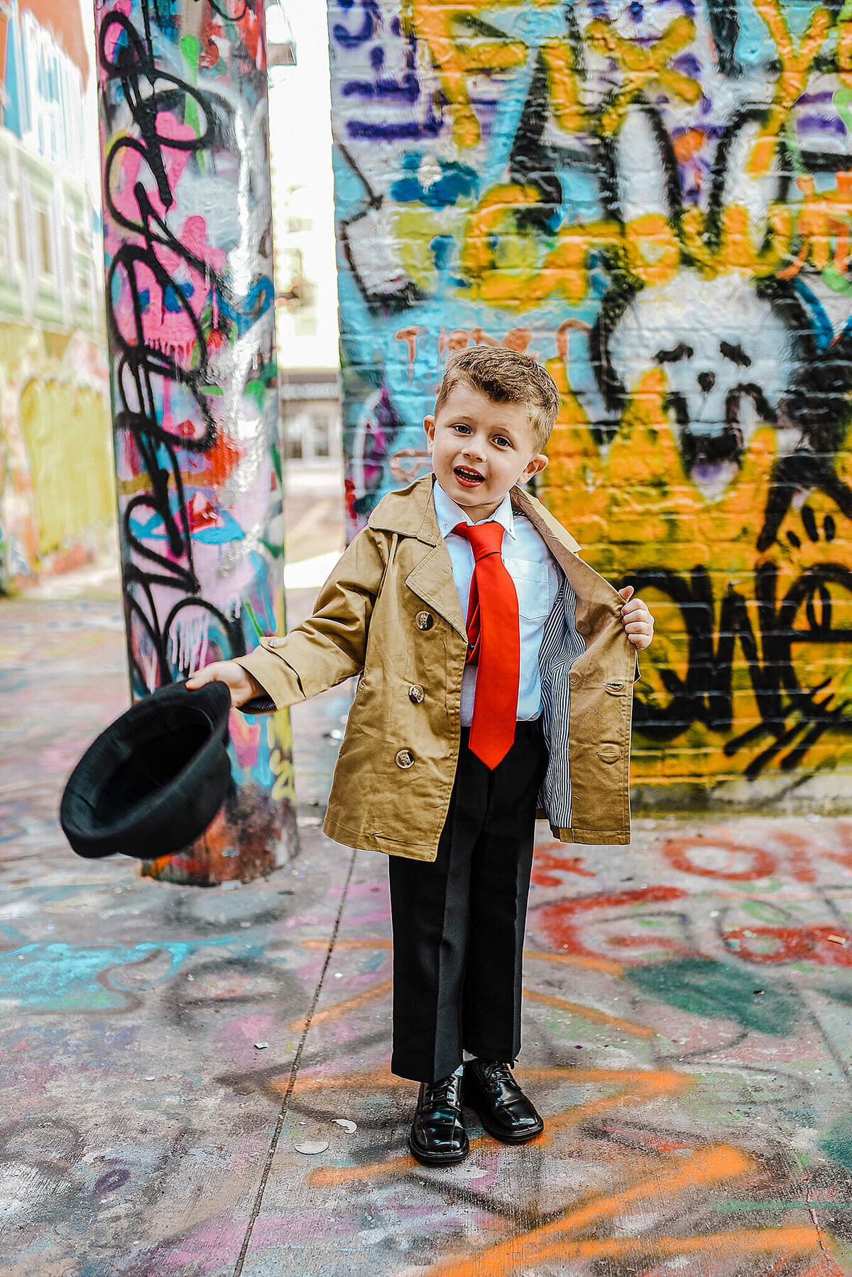 Little boy holding a black fedora wearing a trench coat and a red tie in graffiti alley near MICA in Baltimore Maryland