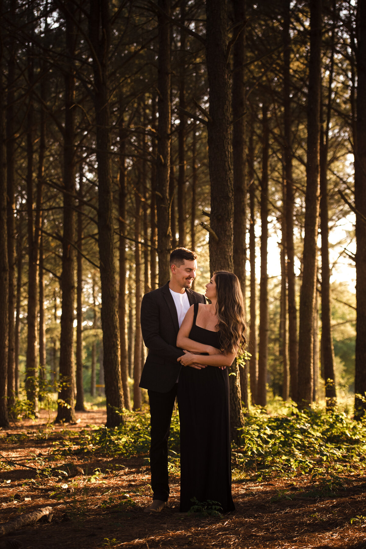 Engaged-couple-in-black-clothing-the-man-standing-behind-the-woman-with-his-arms-around-her-both-looking-at-one-another-smiling-posed-amid-trees-at-Frank-Liske-Park