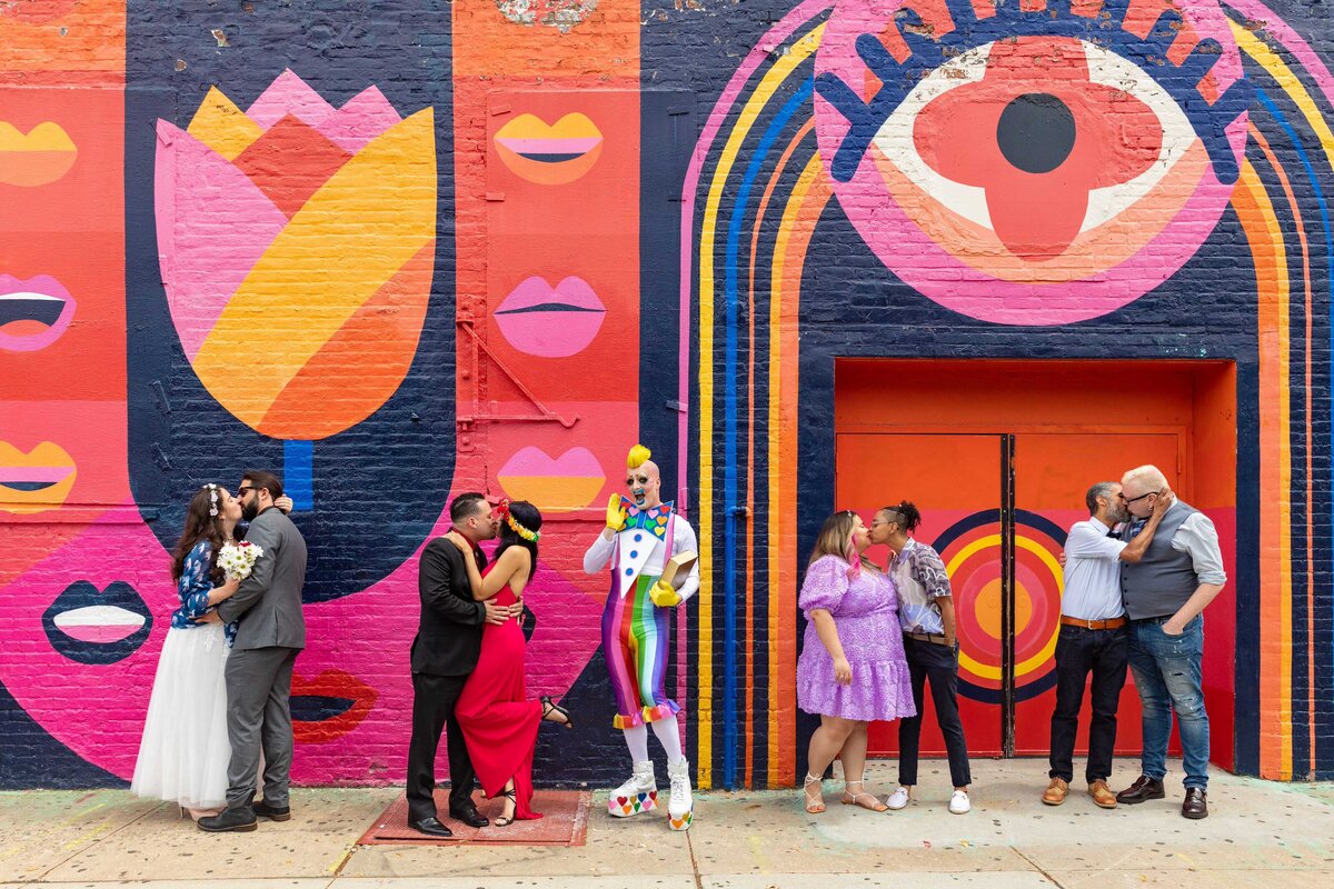 Multiples couples standing in front of a bright mural kissing.